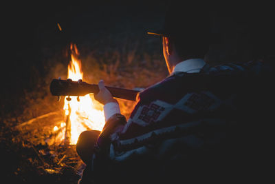 Rear view of man with guitar sitting by bonfire in forest