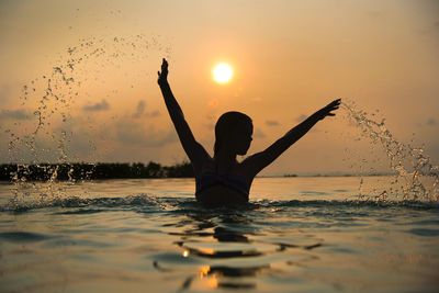 Silhouette teenage girl with arms raised splashing water in sea against sky during sunset