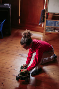 From above side view of ethnic child in casual clothes sitting on wooden floor and playing with toy car at home