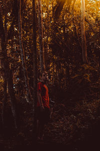 Young man standing against trees in forest during sunset