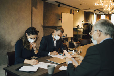 Male and female colleagues writing on paper in board room during covid-19