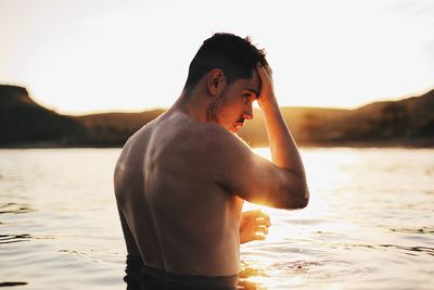 Full length of shirtless man in lake against sky during sunset