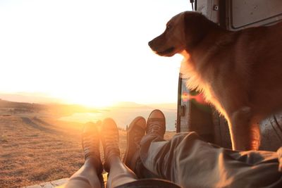 Low section of man standing with dog against sky during sunset