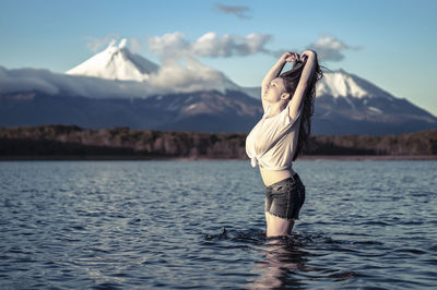 Young woman with arms raised standing in lake against snowcapped mountain