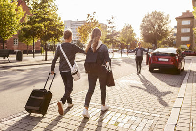 Rear view of siblings with suitcase walking towards smiling mother on roadside