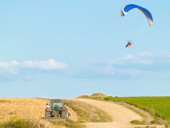 Tractor on field against person paragliding in sky