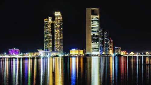 Illuminated modern buildings by river against sky at night