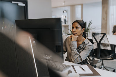 Confident businesswoman with hand on chin using computer while sitting at desk in corporate office