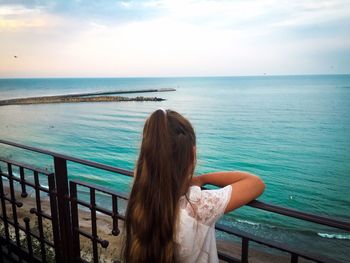 Rear view of girl with long brown hair standing on the bridge and looking at the ocean