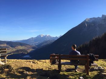 Rear view of mid adult man looking at mountain while sitting on bench against clear blue sky