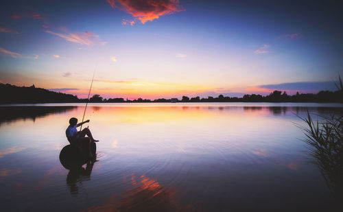 Man fishing in lake against sky during sunset