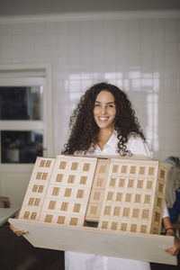 Portrait of smiling female architect holding building model while standing at office