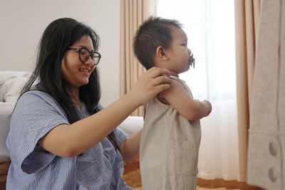 A side view of a young asian mother is dressing her little son. the baby is turned away from his mom