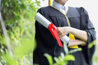 Midsection of young woman in graduation gown with arms crossed standing in park