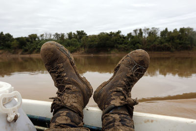 Low section of person wearing messy shoes by river against sky