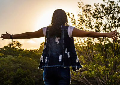 Rear view of woman with arms raised standing against sky during sunset