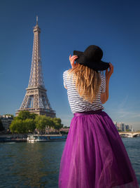 Rear view of woman standing against eiffel tower by river against blue sky