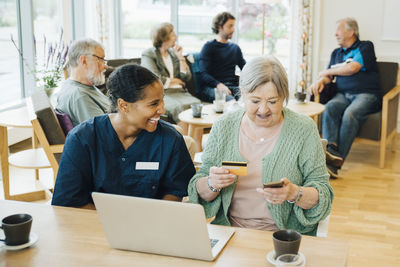 Cheerful female healthcare worker sitting by senior woman doing online shopping with credit card and smart phone in nurs