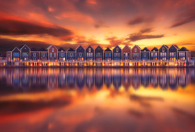 Row of houses reflecting in river against sky during sunset