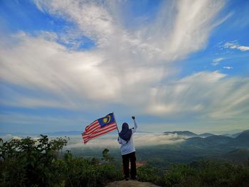 Rear view of woman holding flag standing on mountain against sky