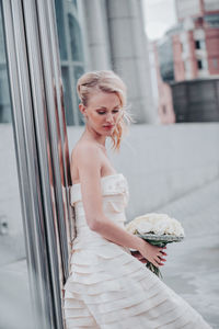 Beautiful bride with bouquet against wall in city