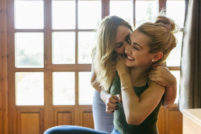 Affectionate mother kissing blond teenage daughter on cheek at home