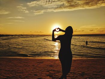 Silhouette woman forming heart shape at beach during sunset