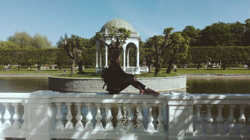 Side view of young woman sitting on retaining wall by pond against sky