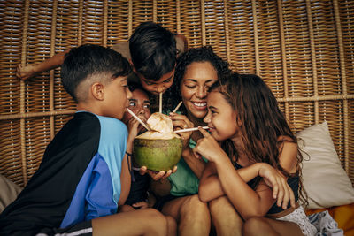 Happy siblings drinking coconut water with mother inside wicker hut at beach