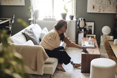 Side view of woman with disability picking up coffee cup from table while sitting on sofa at home