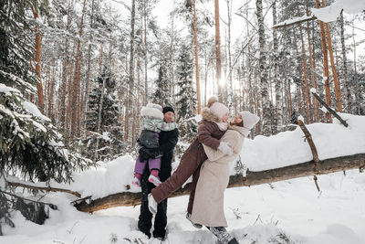 Mom and dad hold their daughters in their arms and have fun in the snowy sunny forest. 