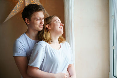 Shot of happy young couple standing together by window while looking away.