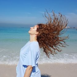 Woman with tousled hair standing at beach against blue sky