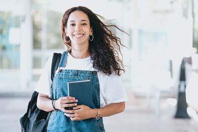 Portrait of young woman holding book while standing in city