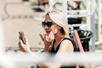 A young woman takes a selfie sitting on a park bench, adjusts her makeup and hairstyle