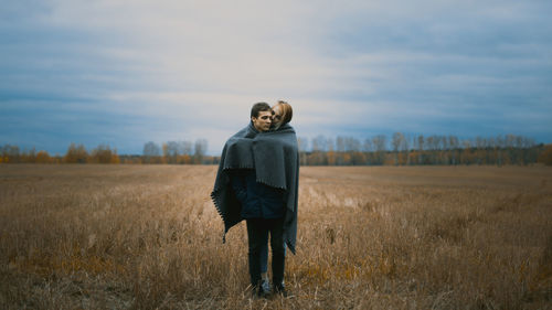 Couple wrapped in blanket on field against cloudy sky