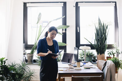 Confident female architect writing in diary while standing by desk in home office