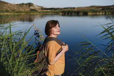 Woman with eyes closed holding backpack while standing near lake