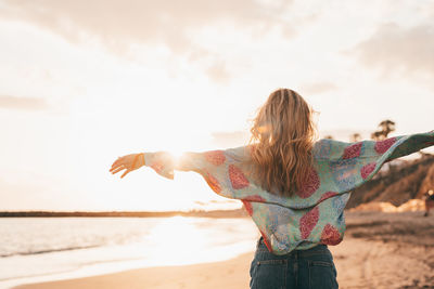 Rear view of woman with arms raised standing at beach against sky