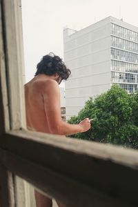 Shirtless man with cigarette standing in balcony seen through window