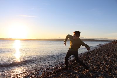 Full length of man throwing pebbles in sea while standing on shore during sunset