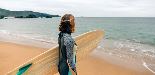 Surfer woman with wetsuit carrying surfboard looking to the sea at the beach