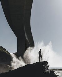 Man standing on top of rock against splashing waves under sea cliff bridge