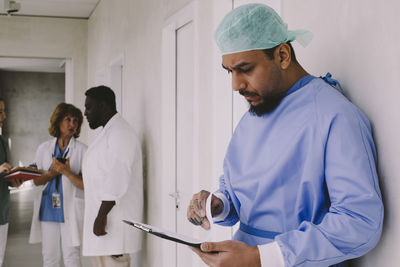 Male surgeon analyzing records while standing near colleagues discussing in corridor at hospital