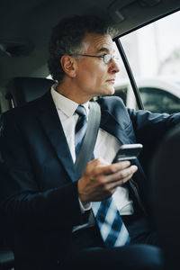 Businessman with smart phone looking through window while sitting in taxi