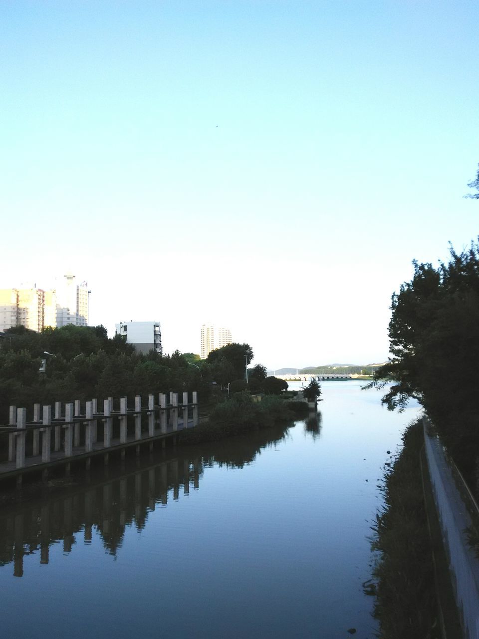 clear sky, water, copy space, built structure, reflection, architecture, building exterior, waterfront, river, lake, blue, tree, tranquility, outdoors, nature, standing water, no people, day, tranquil scene, sunlight