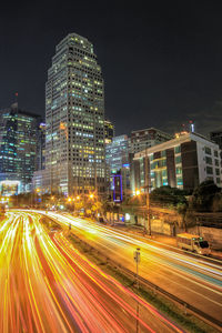 Light trails on city street by buildings against sky at night