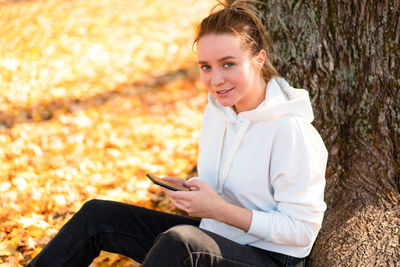 Portrait of smiling woman using smart phone while sitting by tree in park
