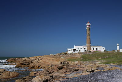 Lighthouse at rocky shore against clear blue sky