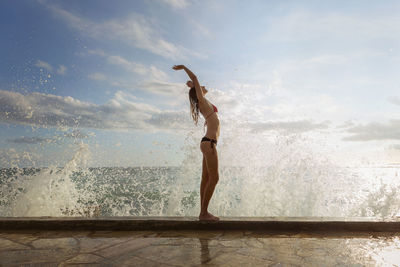 Woman standing with arms raised on wet shore against sky